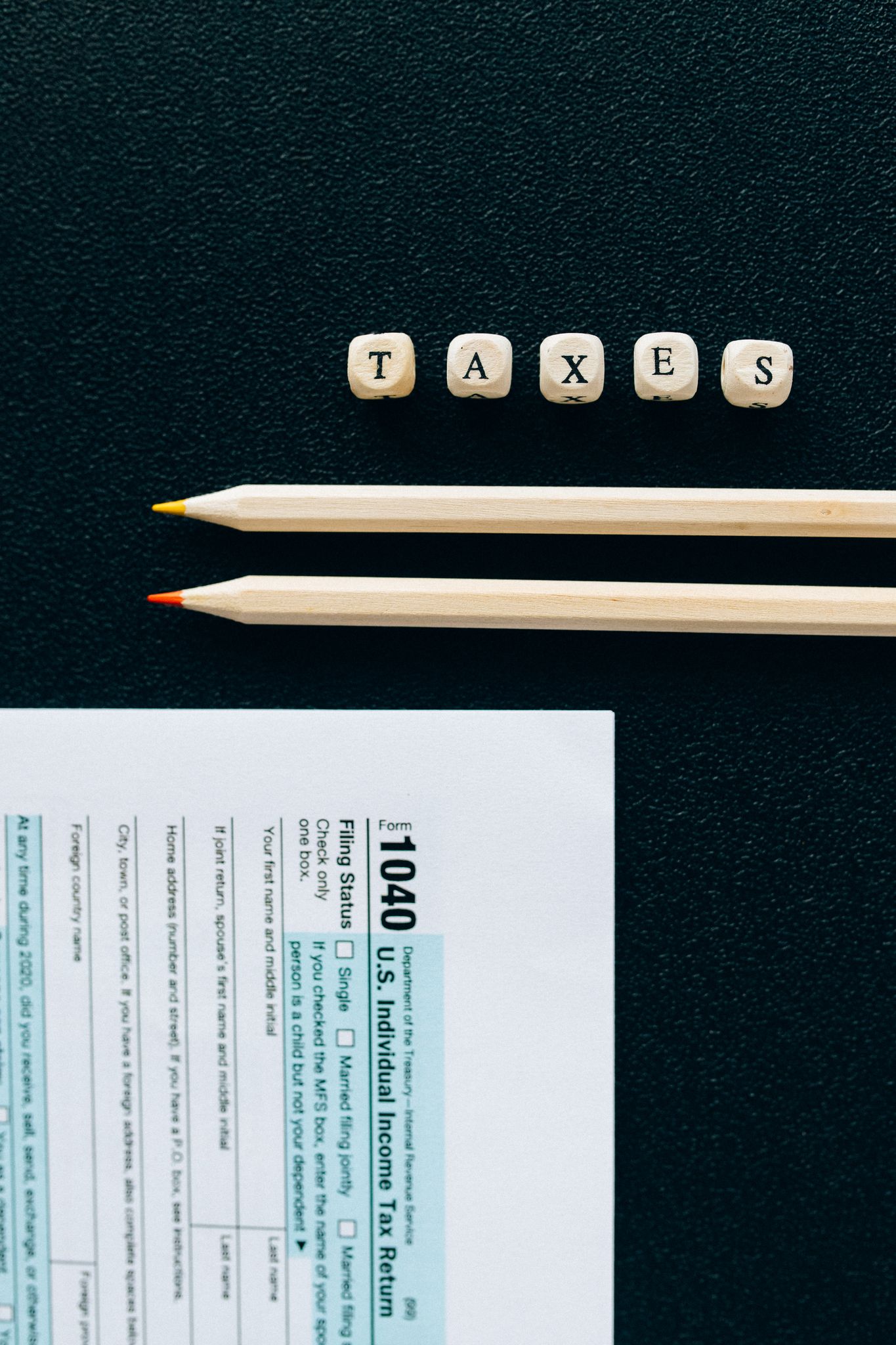 Wooden pencils and 1040 tax form on black background with tiles spelling 'TAXES'.